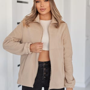 May include: A beige fleece zip-up jacket with a high collar and elasticized cuffs, worn over a white cropped top and black high-waisted button-fly jeans, paired with a beige patterned baseball cap and a matching beige patterned handbag.