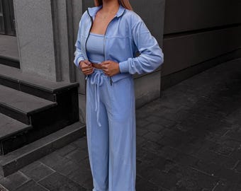 May include: A light blue three-piece set consisting of a cropped zip-up track jacket with a high collar, a matching bandeau top, and high-waisted wide-leg trousers featuring a drawstring waist and vertical seam detailing.