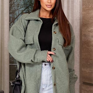 Könnte beinhalten: Eine Frau trägt eine salbeigrüne, geknöpfte Jacke mit zwei Vordertaschen. Die Jacke wird mit einem schwarzen Oberteil und hellblauen Jeans kombiniert. Eine schwarze Handtasche hängt an ihrer Schulter.