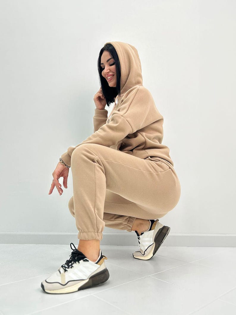 May include: A beige hooded sweatshirt and matching sweatpants set. The outfit is made of a soft material. The person is wearing white and black sneakers with a black lace design. The background is a plain white wall.