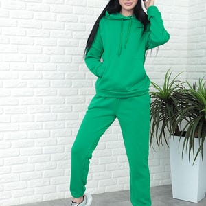May include: A green tracksuit consisting of a hooded sweatshirt and matching sweatpants. The sweatshirt has a drawstring hood and a front pocket. The sweatpants have an elastic waistband and cuffs. The outfit is worn with gray and white sneakers.