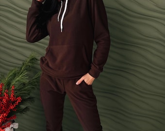 May include: A brown fleece hoodie and matching sweatpants set. The hoodie has a drawstring and a front pocket. The model is wearing black sneakers. The background is a textured green wall.