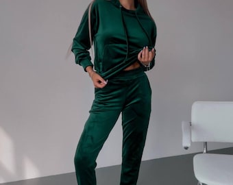 May include: A dark green velvet tracksuit consisting of a hooded sweatshirt and matching pants. The outfit is worn with white sneakers. The tracksuit has a drawstring on the hood and a relaxed fit.