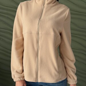 May include: A beige fleece jacket featuring a high collar, a full-length front zipper, and long sleeves with elasticized cuffs. The garment has a horizontal seam across the chest and is paired with blue denim jeans, creating a simple, functional look.
