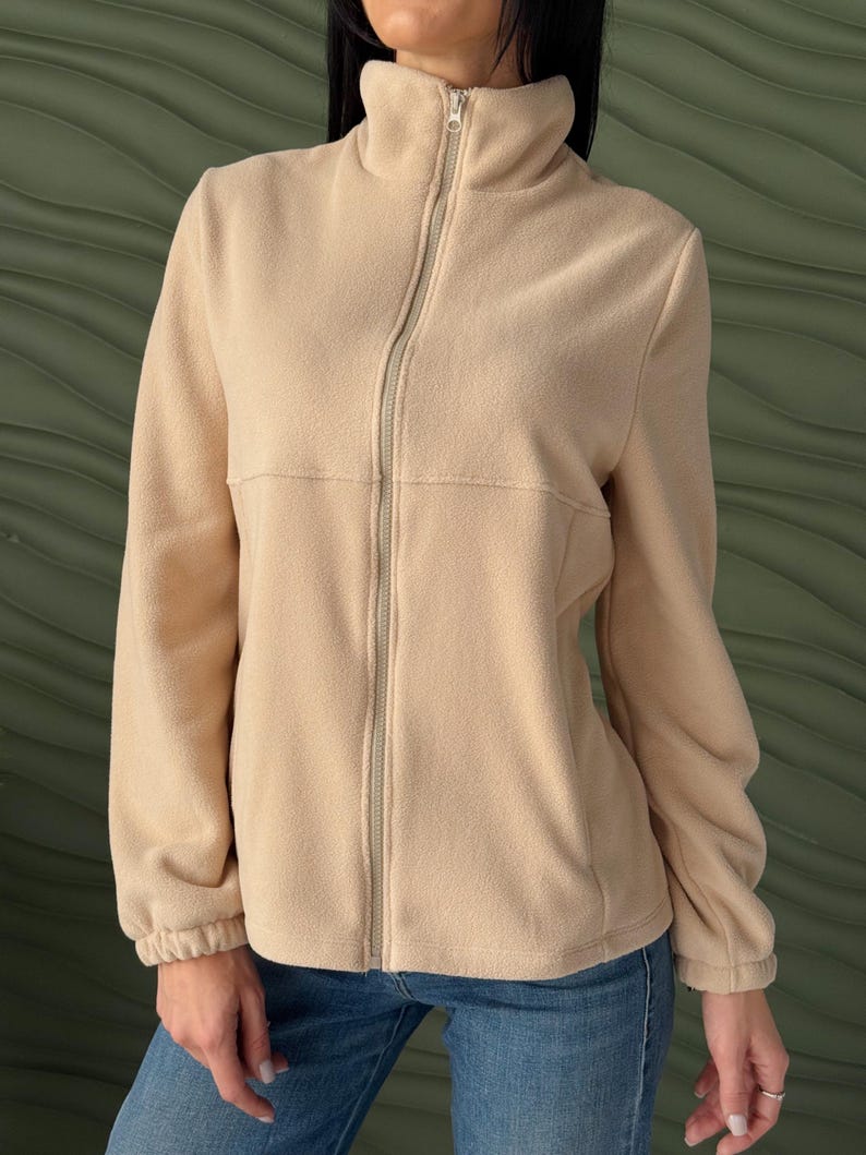 May include: A beige fleece jacket featuring a high collar, a full-length front zipper, and long sleeves with elasticized cuffs. The garment has a horizontal seam across the chest and is paired with blue denim jeans, creating a simple, functional look.