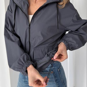Puede incluir: Chaqueta con capucha gris carbón con cremallera negra y cordones. La chaqueta tiene cintura ajustable con cordón y puños elásticos. La modelo lleva vaqueros azules.