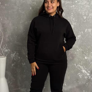 May include: A black hoodie with a drawstring and a front pocket, paired with matching black sweatpants. The outfit is made of a soft material and is suitable for casual wear. The model is smiling.