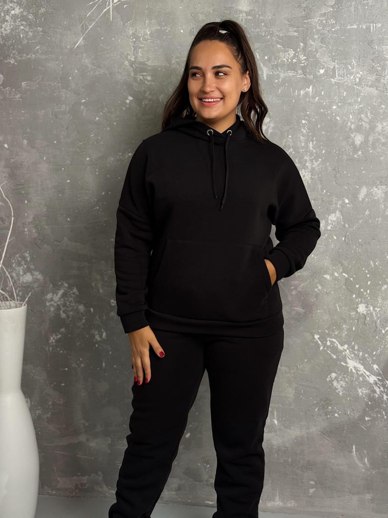 May include: A black hoodie with a drawstring and a front pocket, paired with matching black sweatpants. The outfit is made of a soft material and is suitable for casual wear. The model is smiling.