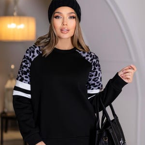 Pode incluir: Camisola preta com mangas estampadas de leopardo e riscas brancas. A modelo usa um gorro preto e uma mala tote preta. Os jeans são azuis.