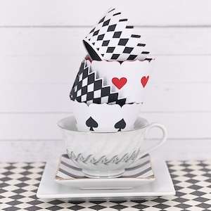 Puede incluir: Una pila de vasos de papel decorativos con patrones de diamantes, corazones y picas en blanco y negro, encima de una taza y un platillo blancos. La taza y el platillo están sobre un plato cuadrado con un borde a rayas blanco y negro, sobre un mantel a cuadros.