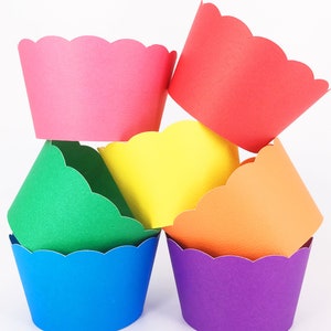Op de afbeelding: Een stapel kleurrijke cupcakevormpjes in roze, rood, groen, geel, oranje, blauw en paars. Elk vormpje heeft een geschulpte rand. De vormpjes zijn in een aantrekkelijke, gestapelde formatie geplaatst tegen een witte achtergrond.