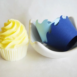 May include: A decorative cupcake with yellow frosting and a white paper liner sits next to a white ceramic dish holding blue paper crown cutouts. The cupcake has a swirl design.