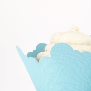 May include: A close-up of a cupcake in a light blue scalloped paper cup. The cupcake has white frosting with dark specks. The background is plain white, highlighting the dessert.