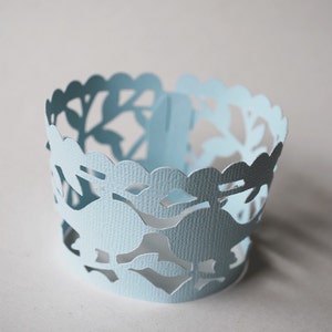 Puede incluir: Un envoltorio de papel azul claro para cupcakes con un borde festoneado y intrincados recortes de hojas. El envoltorio cilíndrico tiene una superficie texturizada, perfecto para añadir un toque decorativo a los productos horneados.