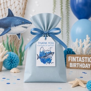 May include: A light blue gift bag tied with a blue ribbon and a tag that says "Thank You for making my birthday" with a shark illustration. The bag is decorated with a shark theme, including a shark cutout and balloons.