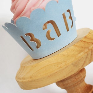 May include: A cupcake with pink frosting sits in a light blue paper cup that reads "BAB" in cut-out letters. The cupcake rests on a wooden cake stand with a round top and a pedestal base. The background is white.