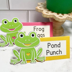 May include: Green frog-shaped paper cutouts with white outlines and heart details. The frogs sit in front of colorful signs that read "Frog Eggs" and "Pond Punch". The signs are pink and yellow with white text.