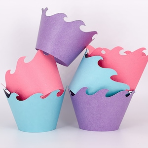 May include: A stack of colorful cupcake liners in shades of purple, pink, and blue. Each liner has a wavy, scalloped edge. The liners are arranged in a playful, overlapping manner against a white background.
