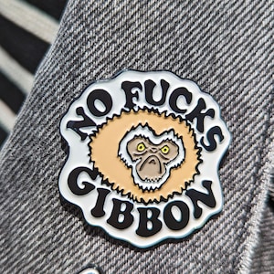 May include: A white and black enamel pin with the text "NO FUCKS GIBBON" and a cartoon gibbon face. The pin is attached to a gray denim jacket.