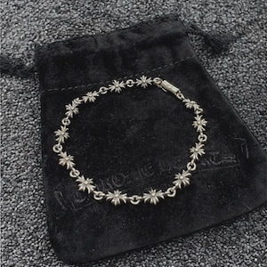 Puede incluir: Un brazalete plateado con abalorios en forma de estrella. Se muestra sobre una bolsa de terciopelo negro. El fondo es una superficie gris texturizada.