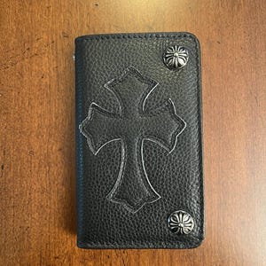 May include: Black leather wallet with a large cross design and silver-colored studs. The wallet has a textured surface and a rectangular shape. The cross is outlined in a lighter color, creating a contrast against the black leather.