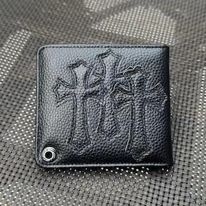 Peut inclure: Portefeuille en cuir noir avec un motif de croix en relief. Le portefeuille présente une surface texturée et un détail d'œillet en métal. Le design comprend trois croix proéminentes, ajoutant une esthétique unique.