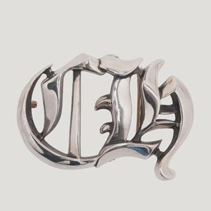 Puede incluir: Hebilla de cinturón plateada con un diseño ornamentado y estilizado de la letra "D". La hebilla tiene un acabado pulido y una estética clásica de inspiración gótica. El diseño presenta curvas fluidas y ángulos agudos.