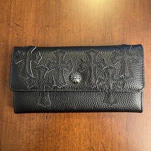 Könnte beinhalten: Schwarze Ledergeldbörse mit strukturierter Oberfläche und silberfarbenem Knopfverschluss. Die Geldbörse hat ein geprägtes Design auf der Vorderseite und einen Klappverschluss. Die Geldbörse ist rechteckig.