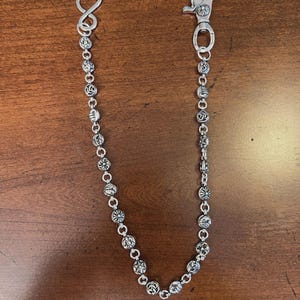 May include: Silver-toned wallet chain with decorative ball links and a lobster clasp. The chain features various embossed designs on the ball links and an infinity loop at the other end. The chain is displayed on a brown wooden surface.