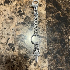 Puede incluir: Llavero plateado con un colgante decorativo de daga y una cadena de eslabones. El llavero incluye un cierre para sujetar y un anillo para llaves. El colgante de la daga tiene detalles intrincados.
