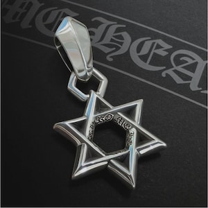 May include: A silver pendant in the shape of a Star of David. The pendant has a polished finish and a bail for attaching to a chain. The star has an inscription around the center. The background is dark with a decorative pattern.