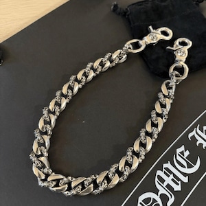 Op de afbeelding: Een zilverkleurige ketting met een gourmette-schakelontwerp. De ketting heeft decoratieve kruisaccenten op elke schakel en is beveiligd met twee karabijnsluitingen. De ketting wordt weergegeven op een donkere ondergrond.