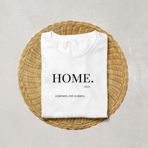 Puede incluir: Camiseta blanca con la palabra "HOME." en negrita, en negro. Debajo de la palabra "HOME" está la palabra "sustantivo" en una fuente más pequeña. Debajo, la frase "una persona, no un lugar." está impresa en una fuente más pequeña. La camiseta está sobre una superficie redonda, tejida, de color marrón claro.