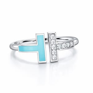May include: A silver ring with a modern design. The ring features a light blue enamel section and a section adorned with clear crystals. The ring is set against a white background, highlighting its elegant details.