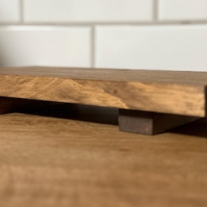 Könnte beinhalten: Ein rechteckiger Holzaufsteller mit dunkler Beize. Der Aufsteller wird von zwei rechteckigen Blöcken getragen. Die Holzmaserung ist sichtbar. Dieser Artikel eignet sich zur Präsentation kleiner Objekte.