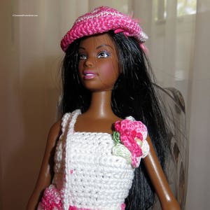 May include: A black doll with long black hair wearing a white and pink crocheted dress and a pink crocheted hat with a white stripe.