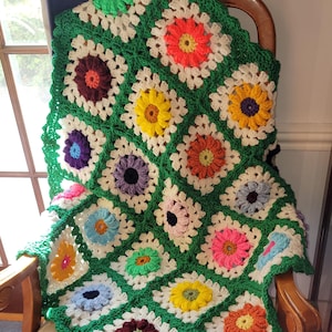 May include: A crocheted blanket with a green background and colorful flower designs. The blanket is folded and draped over a wooden chair.