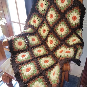 May include: A crocheted blanket with a brown, green, and white daisy pattern. The blanket is draped over a wooden chair.