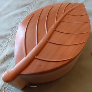 May include: A wooden box shaped like a leaf, with a light brown finish and detailed carvings. The box is closed and the lid is slightly ajar.
