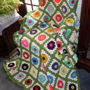 May include: A handmade crochet blanket draped over a chair. The blanket features a green background with colorful floral squares in shades of red, pink, yellow, blue, and purple. The blanket's edge is scalloped.