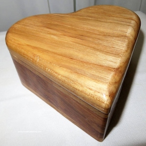 May include: A heart-shaped wooden box with a light brown lid and a darker brown base. The box is made of wood and has a smooth, polished finish. The lid is curved and fits snugly on the base. The box is likely used for storing small items.