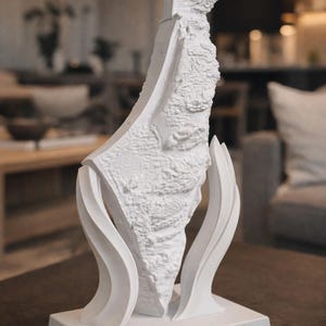 Könnte beinhalten: Eine weiße Skulptur einer Landmasse, möglicherweise eine Karte, mit strukturierten Details und einem stilisierten Sockel. Die Skulptur wird von geschwungenen, flügelartigen Elementen getragen. Das Stück steht auf einer dunklen Oberfläche mit verschwommenem Hintergrund.