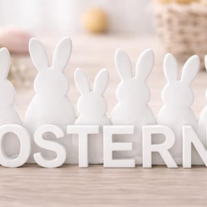 Könnte beinhalten: Weiße Holzfiguren von Osterhasen, angeordnet in einer Reihe, mit dem Wort "OSTERN" in weißen Buchstaben davor. Die Hasen haben lange Ohren und sind unterschiedlich groß. Der Hintergrund ist verschwommen, was eine weiche, natürliche Umgebung suggeriert.