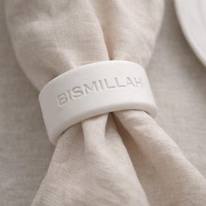 Könnte beinhalten: Ein weißer Keramik-Serviettenring mit dem geprägten Wort "BISMILLAH". Er umschließt eine beige Leinen-Serviette. Im Hintergrund sind ein weißer Teller und ein goldenes Besteck zu sehen. Geeignet für Tischdekorationen.