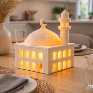 Könnte beinhalten: Eine weiße, moscheeförmige Lampe mit einem Halbmond. Die Lampe hat gewölbte Fenster, die ein warmes Licht ausstrahlen. Sie steht auf einem Holztisch und erzeugt eine warme Atmosphäre. Die Lampe ist ca. 15 cm lang.
