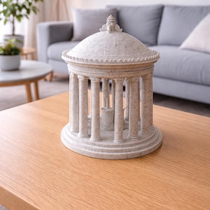 Pode incluir: Um templo em miniatura detalhado, branco sujo, com telhado abobadado e colunas. A peça decorativa está sobre uma mesa de madeira castanha clara. O fundo inclui um sofá cinzento, uma pequena planta em vaso e uma lâmpada.