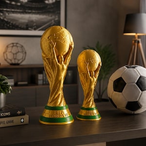 Könnte beinhalten: Zwei goldfarbene FIFA-Weltmeisterschaftspokal-Nachbildungen in verschiedenen Größen, jeweils mit einer Figur, die einen Globus auf einem grünen Sockel mit der Aufschrift FIFA WORLD CUP hält, neben einem klassischen Fußball.