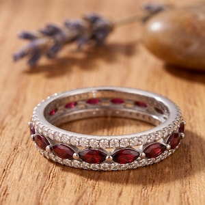 May include: A silver band ring featuring a central row of marquise-cut deep red gemstones flanked by two rows of small, round, clear white stones. The metal band has a polished finish and includes a visible hallmark stamp on the inner surface.