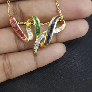 Puede incluir: Un collar de oro con un colgante con diseño de cinta. La cinta está adornada con piedras preciosas rojas, blancas, verdes y azules. El collar es sostenido por una mano sobre un fondo oscuro.