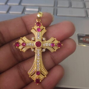 Puede incluir: Colgante dorado en forma de cruz, adornado con piedras preciosas rojas y blancas. La cruz presenta detalles intrincados y un lazo para una cadena. Una joya.